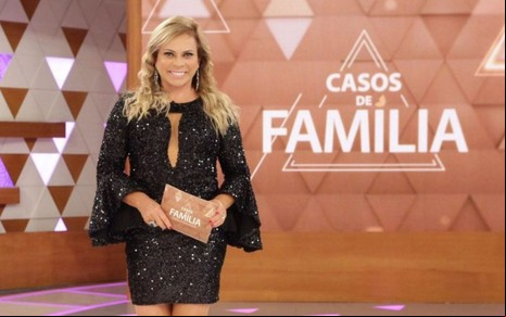 Christina Rocha posando em frente a um telão com o logo do Casos de Família; ela aparece sorridente, com vestido preto de lantejoulas, e segura ficha do programa nas mãos