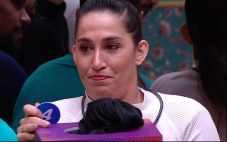 Daniele Hypolito pegando um número de uma urna do BBB 25, sentada no sofá da sala