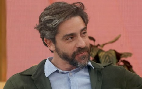 Felipe Abib sentado no sofá do Encontro, usando casaco verde-escuro e camisa azul-claro
