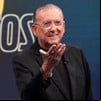 Galvão Bueno no estúdio de Galvão e Amigos; ele aparece todo de preto, estendendo a mão direita, em frente à logo do programa