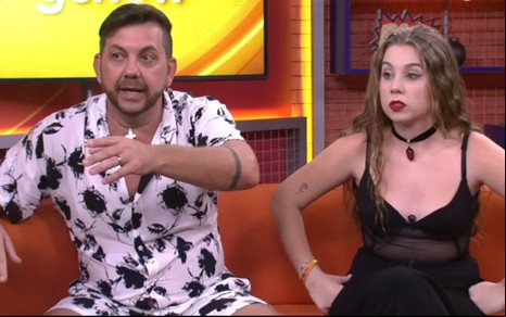Edilberto Simões usa uma camisa estampada e está sentado ao lado da filha, Raissa Simões, que usa um body preto de alcinha; os dois exprimem choque