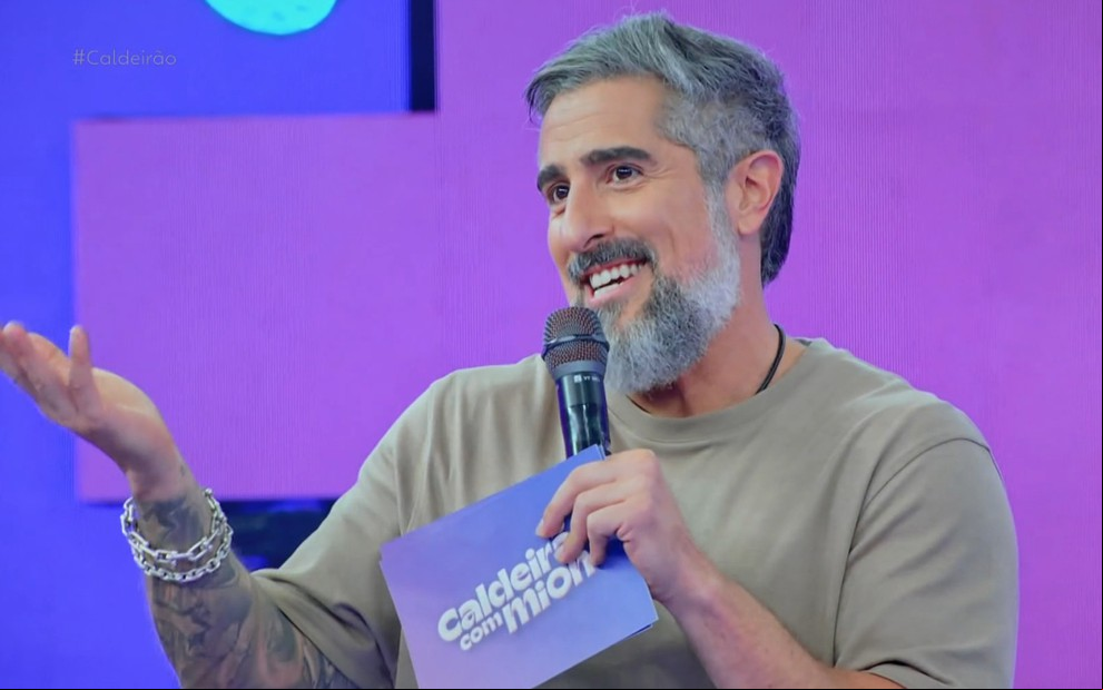 Marcos Mion força sorriso e ergue a mão em gesto constrangedor