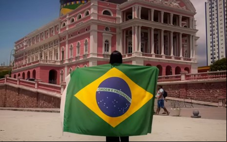 Uma pessoa de costas segura uma bandeira do Brasil aberta diante do Teatro Amazonas, em Manaus, durante o dia