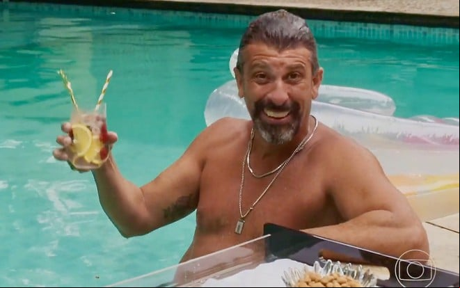 O ator Milhem Cortaz segurando drink dentro da piscina, sorrindo em cena de Volta por Cima