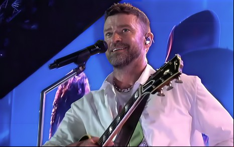 Justin Timberlake tem expressão desconfiada no palco de um show; ele segura um violão