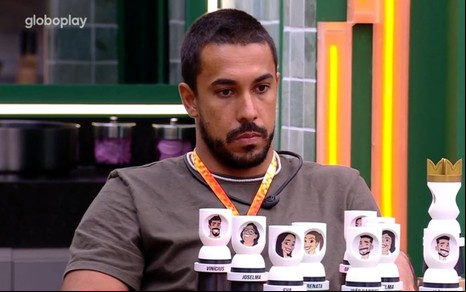Maike Cruz está sentado em uma mesa, com totens dos participantes do BBB 25