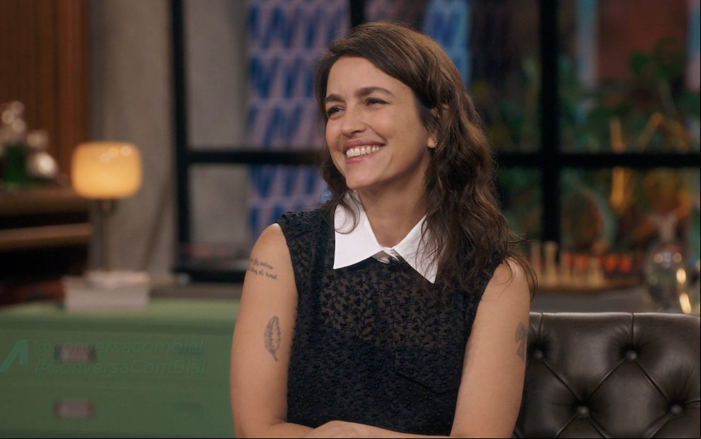 Mulher sorridente em estúdio de TV, com blusa preta rendada e gola branca, sentada em sofá de couro escuro.