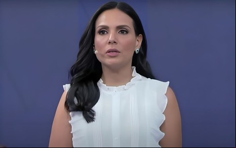 A mulher da imagem parece estar apresentando ou discursando, com expressão séria e cabelos escuros, usando uma blusa branca elegante.