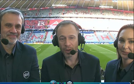 Trio de comentaristas do SBT aparece em cabine de transmissão durante jogo entre PSG e Inter de Milão, com estádio ao fundo.