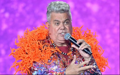 Milton Cunha com plumas laranjas ao redor do corpo segurando um microfone