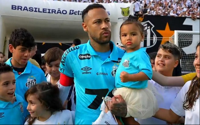 O jogador Neymar, com um uniforme azul do Santos, com a filha Mavie no colo e cercado de crianças na entrada de um campo de futebol