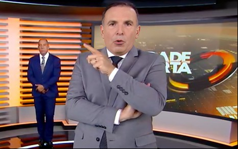 Dois apresentadores em estúdio de telejornal; um ao fundo sério, outro à frente gesticulando com expressão firme.