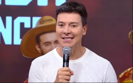 Homem de cabelo curto e camisa branca segura um microfone, com um fundo de palco mostrando um chapéu de cowboy e texto ao fundo.