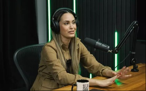 Sarah Andrade está sentada à mesa, com um headphone, e fala em um microfone