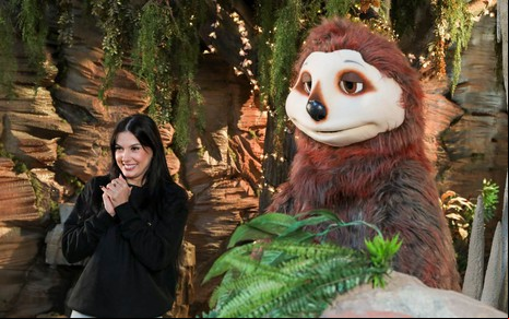 Ana Castela sorri ao lado de um bicho-preguiça de pelúcia gigante em um cenário de caverna