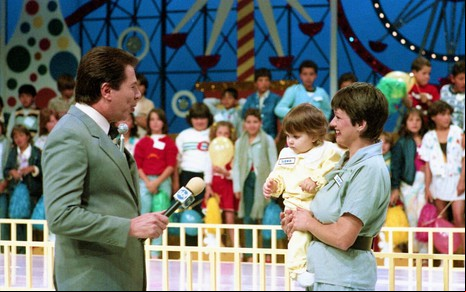 Silvio Santos conversa com uma mãe que segura uma criança no colo no cenário do Domingo no Parque
