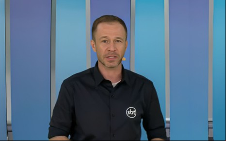 Homem em frente a fundo azul, usando camisa preta com logotipo do SBT. Ele está com microfone de lapela e expressão séria.