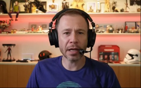 Homem com headset e camisa azul em frente a prateleira com colecionáveis e iluminação quente, parece estar falando ou transmitindo algo.