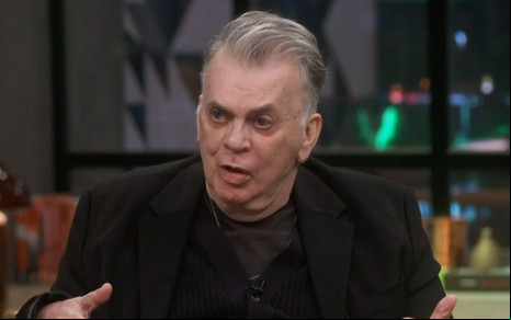 Walcyr Carrasco usando terno preto, sentado no cenário do Conversa com Bial